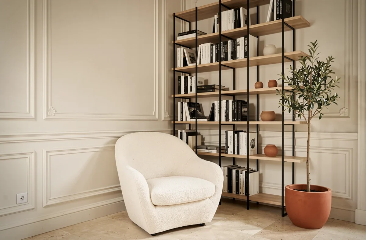 Détail d'angle d'un salon minimaliste parisien avec fauteuil en bouclette blanche, bibliothèque en acier et olivier en pot terracotta